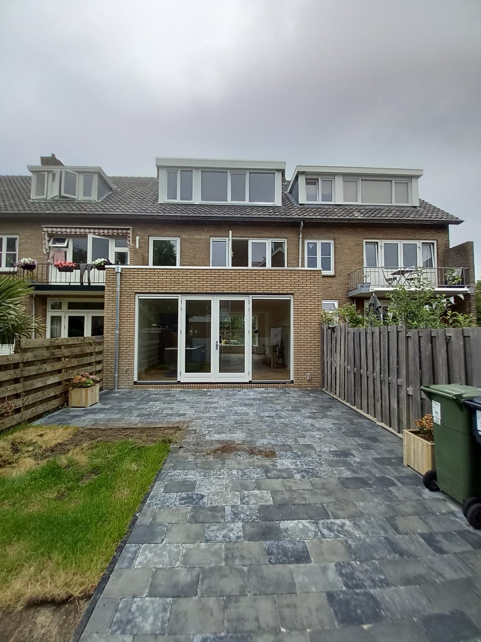 Nowoczesny taras z szarej kostki brukowej przylegający do ceglanego domu z dużymi, przeszklonymi drzwiami tarasowymi. Widoczne drewniane ogrodzenie i fragment trawnika.
