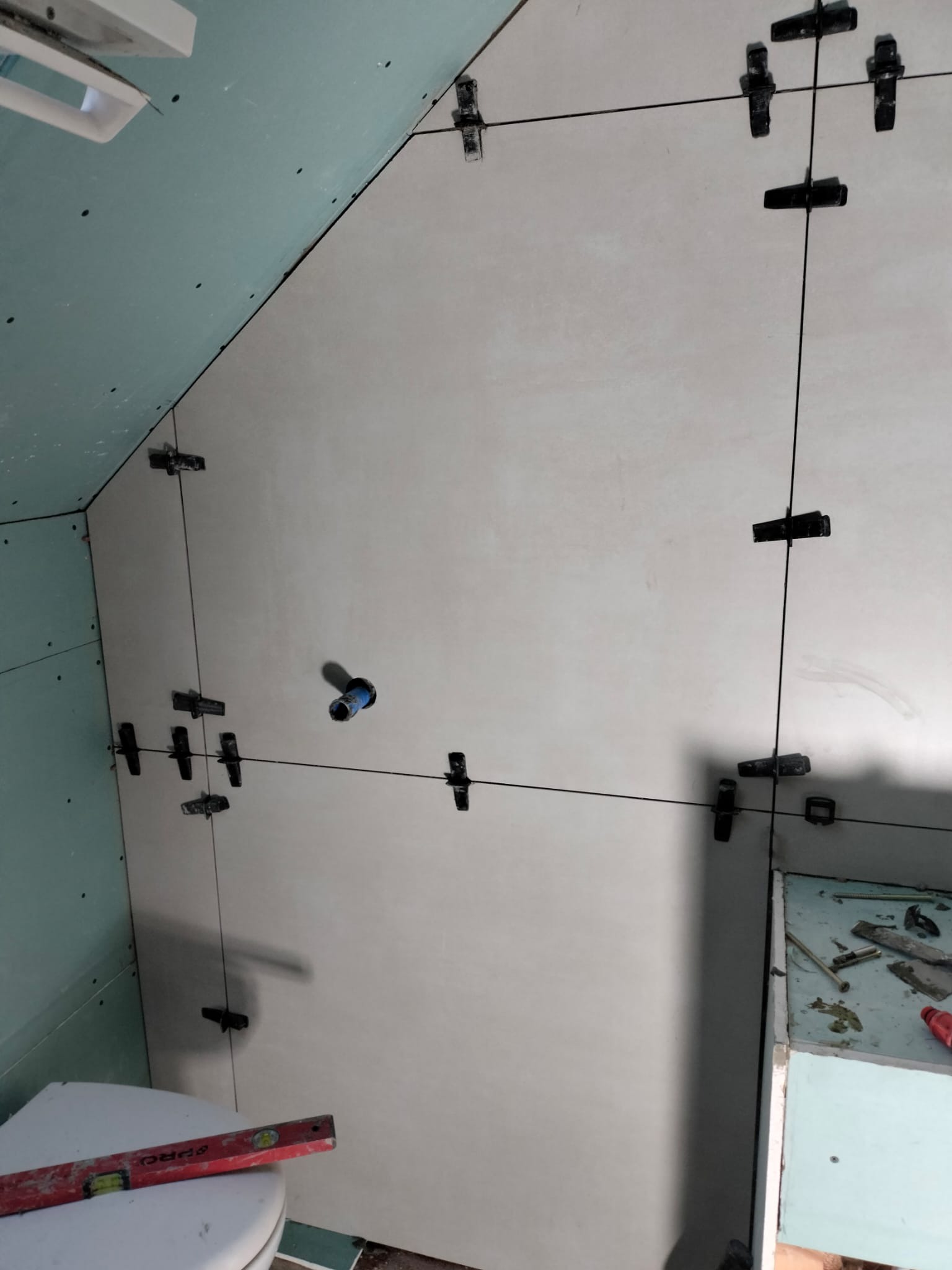 Ściana w łazience z płytami karton-gips i świeżo położonymi, jasnymi płytkami, wyrównywanymi za pomocą klipsów i poziomicy. Widoczny fragment toalety i narzędzia.