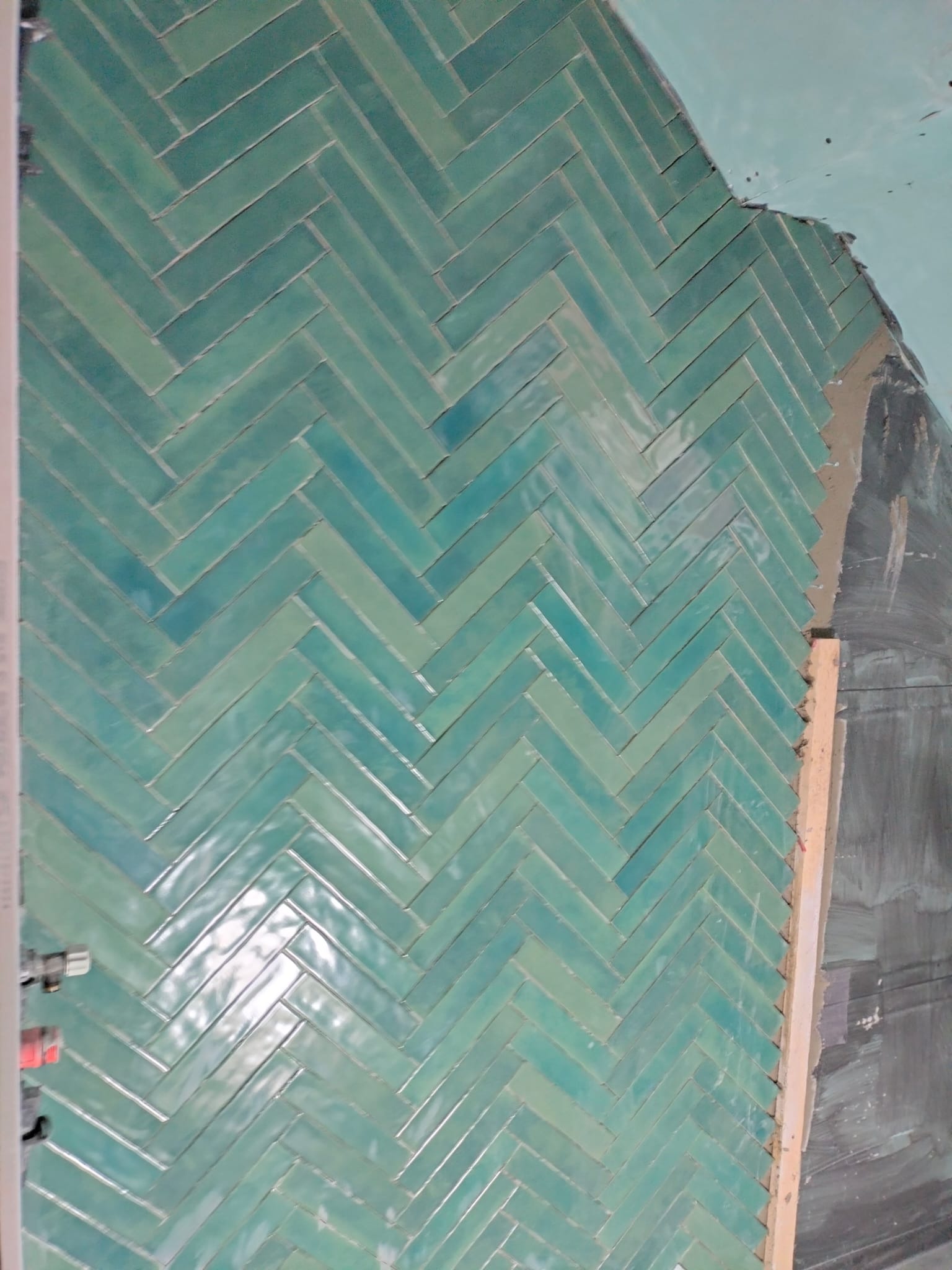 Ułożona, błyszcząca, turkusowa glazura w jodełkę na ścianie łazienki, widoczne fragmenty instalacji i surowego wykończenia. Nietypowy odcień płytek.