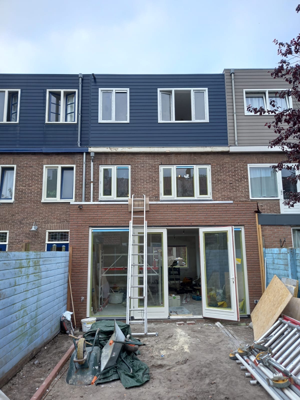 Rozbudowa domu w trakcie: widoczne otwarte drzwi balkonowe, rusztowania wewnątrz, drabina oparta o ścianę. Częściowo wykończona elewacja z drewnianą okładziną.