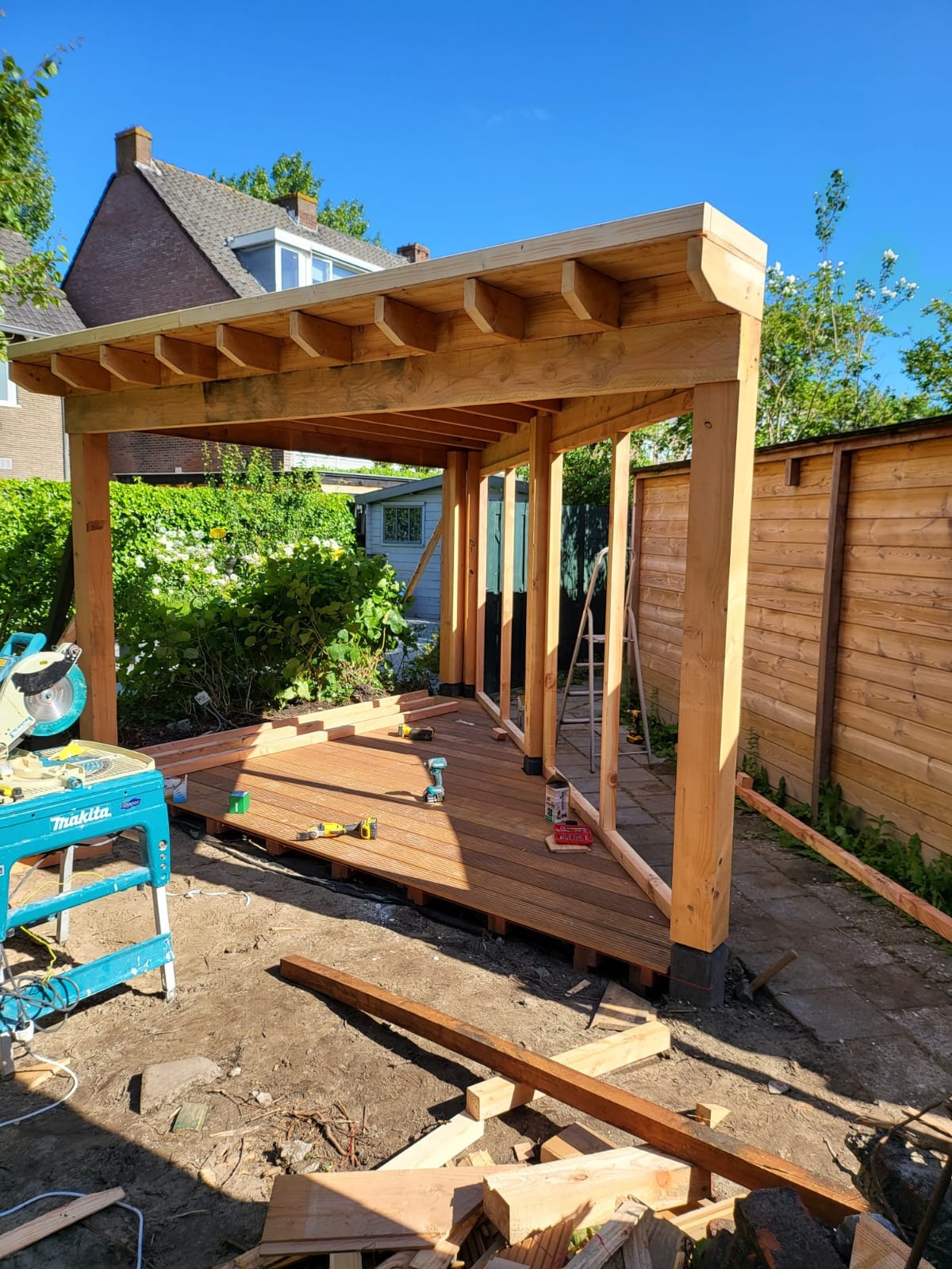 Wznoszenie drewnianej altany w ogrodzie: konstrukcja z belek, desek podłogowych i narzędzi. W tle dom i zieleń. Widoczne etapy budowy, kątowe ułożenie desek podłogowych.