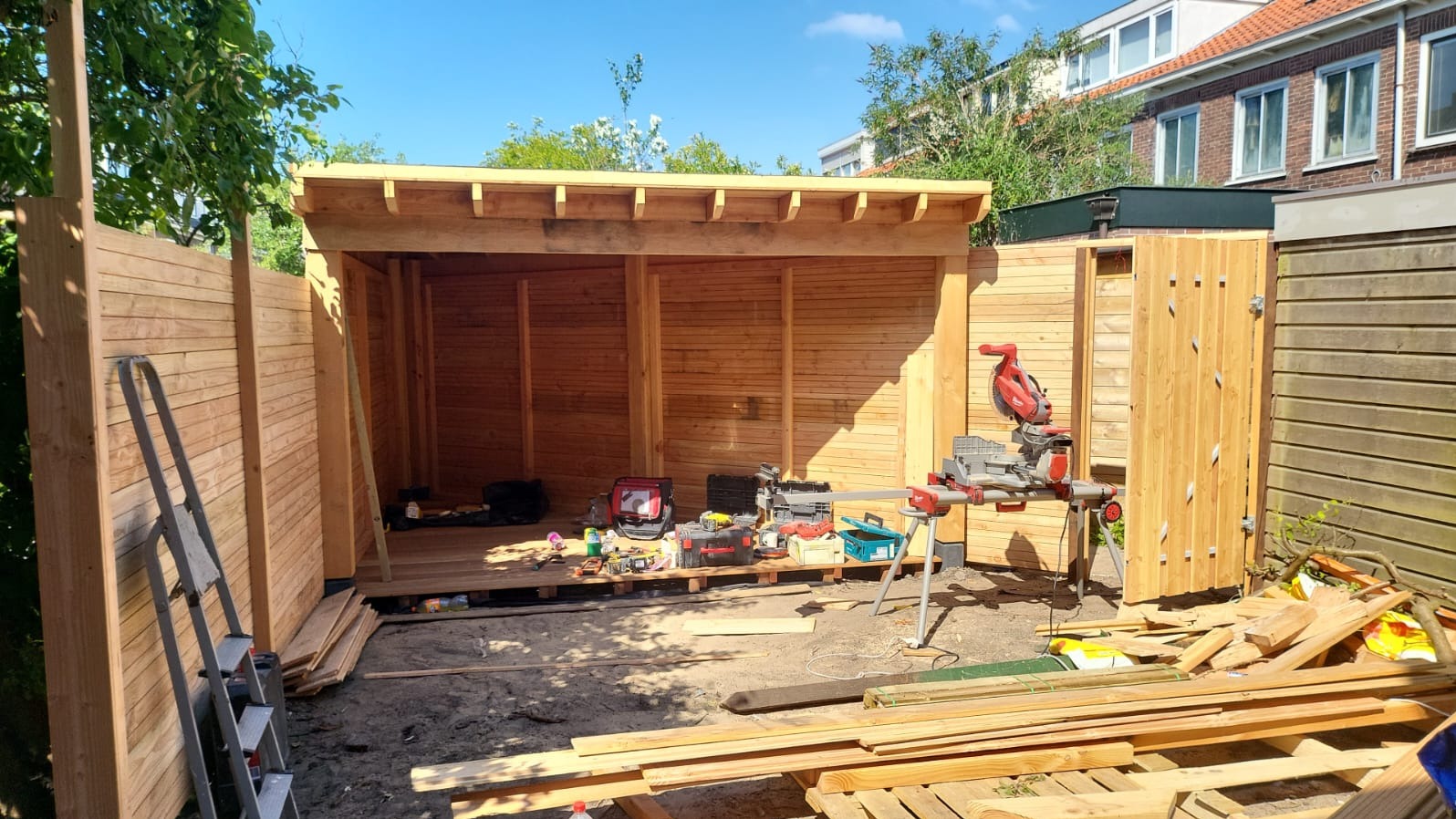 W trakcie budowy drewniana altana z widocznymi narzędziami i materiałami budowlanymi, częściowo ukończone ogrodzenie w tle, słoneczny dzień.