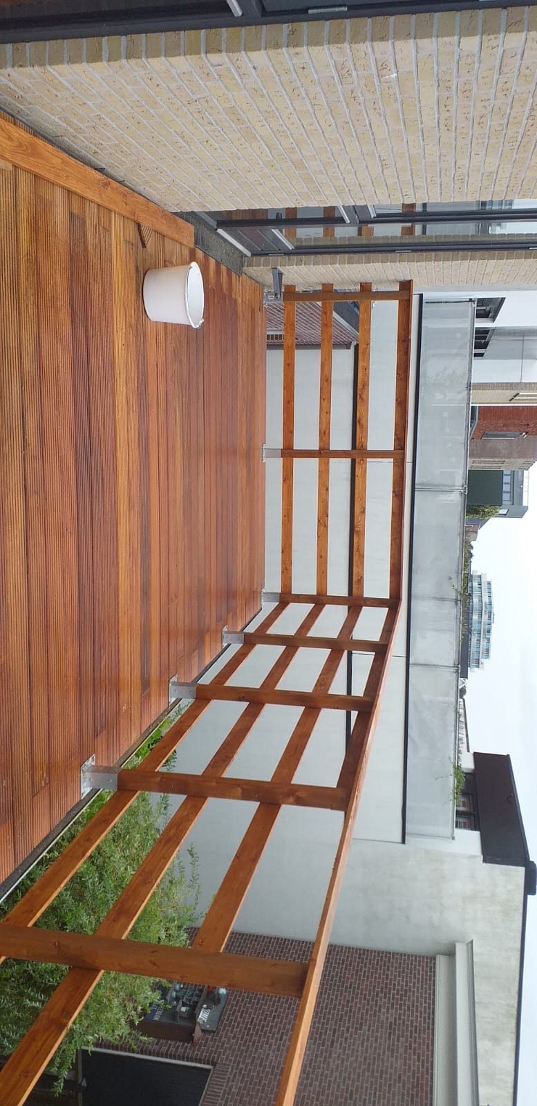 Drewniany taras z balustradą, widok z góry. Deski tarasowe w ciepłym odcieniu brązu, balustrada z drewnianych belek. Białe wiadro przy ścianie. Budynki w tle.