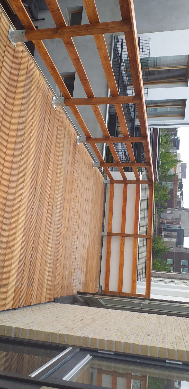 Drewniany taras z pergolą, widok z góry. Deski ułożone pionowo, drewniana konstrukcja pergoli z metalowymi mocowaniami. W tle zabudowa miejska.