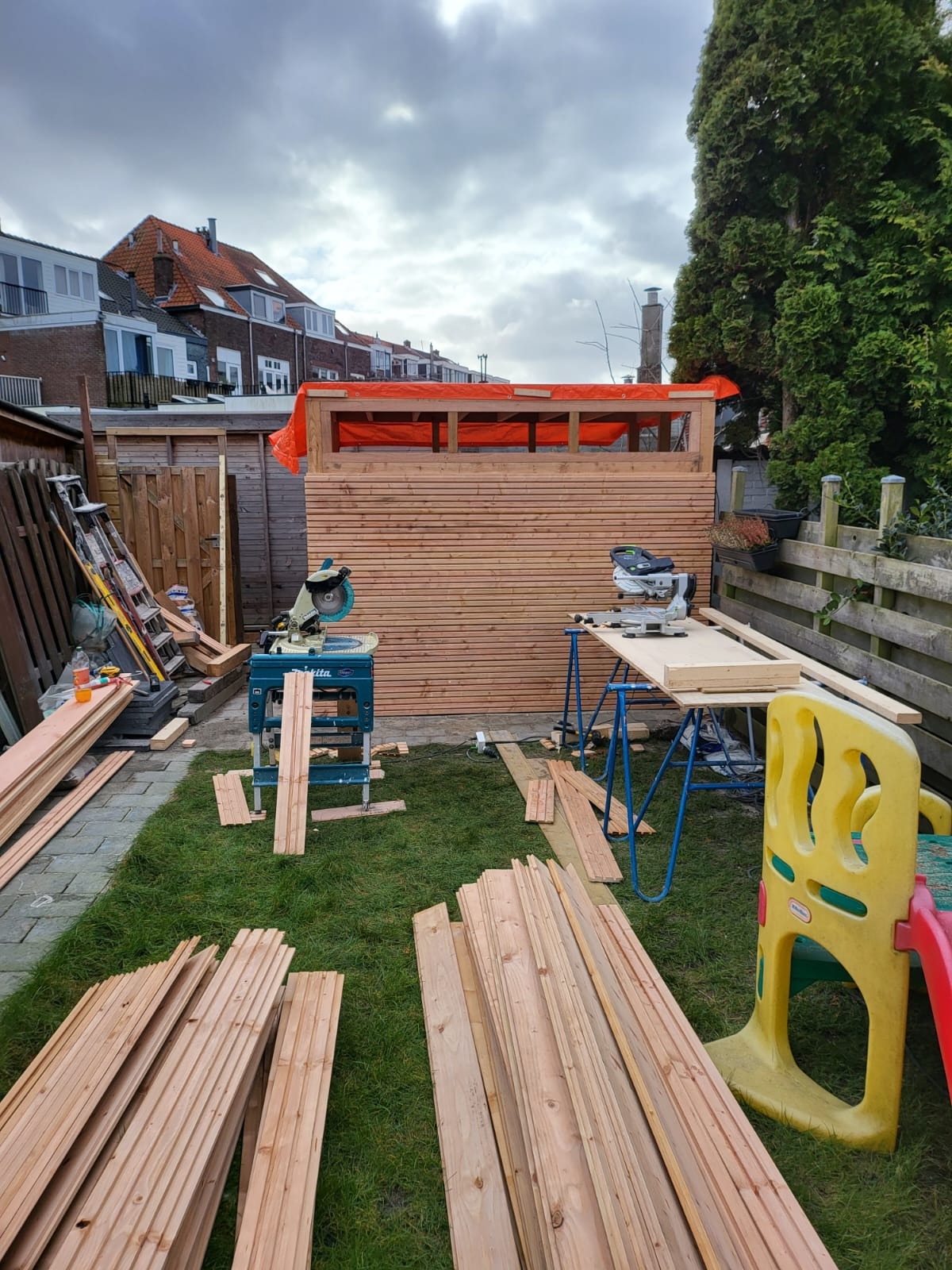 Budowa drewnianej konstrukcji w ogrodzie: deski, piły, trawa. Częściowo ukończona ściana z desek pod pomarańczową plandeką. W tle domy i pochmurne niebo.