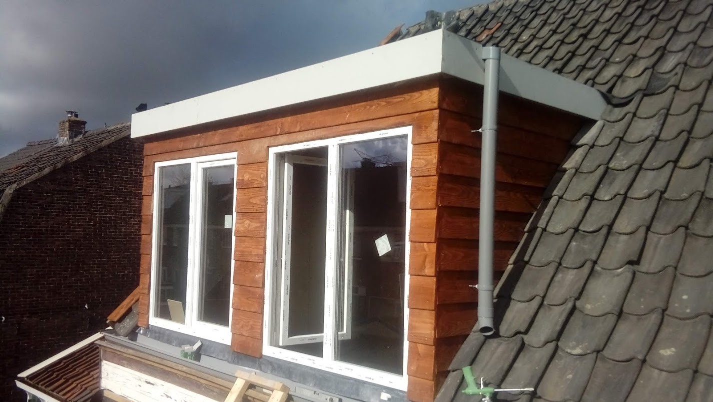 Drewniana nadbudowa lukarny z dwoma oknami PCV, jedno otwarte, na tle dachu z dachówki karpiówki i fragmentu ceglanego budynku. Widoczne narzędzia i pianka montażowa.