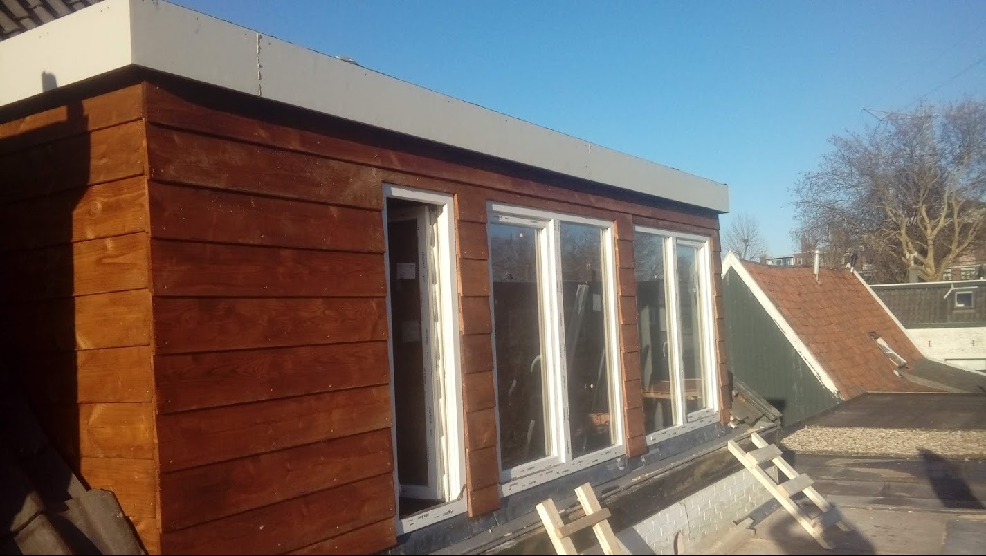 Nowa, drewniana nadbudowa z oknami na dachu, widoczna z zewnątrz w słoneczny dzień. Brązowe deski elewacyjne, białe ramy okienne i drabina oparta o dach.