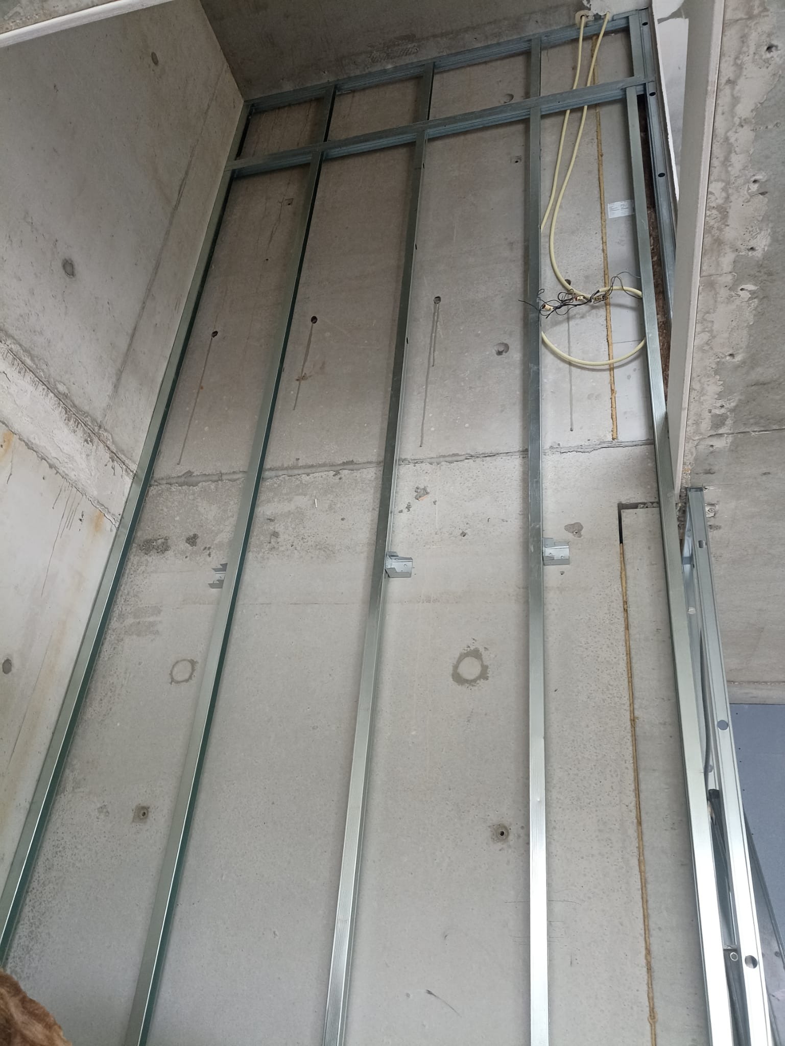 Metalowy stelaż na surowej ścianie, przygotowany pod montaż płyt kartonowo-gipsowych. Widoczne przewody elektryczne. Ujęcie z dołu, podkreślające wysokość pomieszczenia.