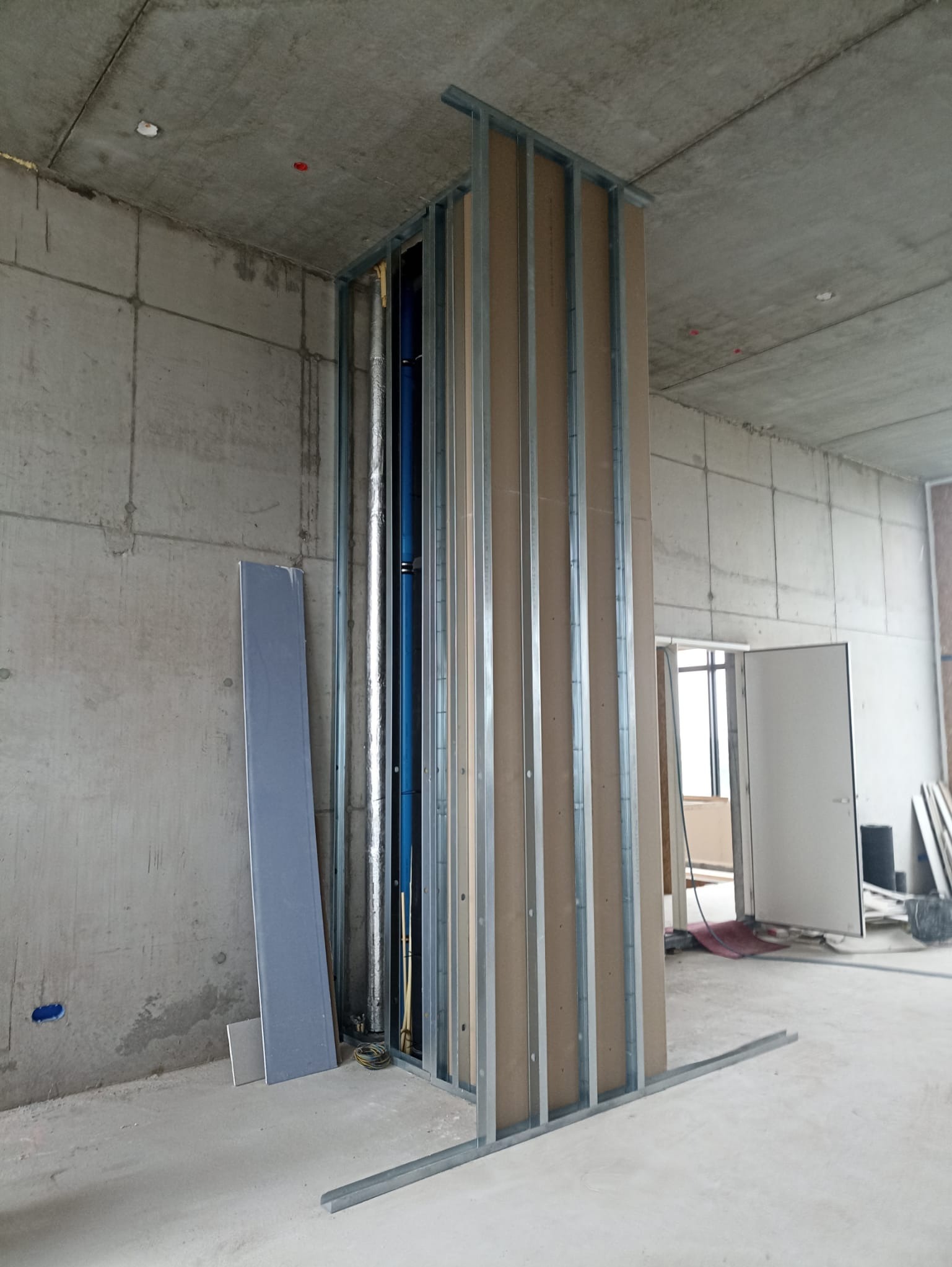 Montaż stelaża z płyt karton-gips na tle surowego betonu. Widoczne pionowe profile i fragmenty płyt. W tle instalacje i otwarte drzwi. Surowy stan wykończenia.