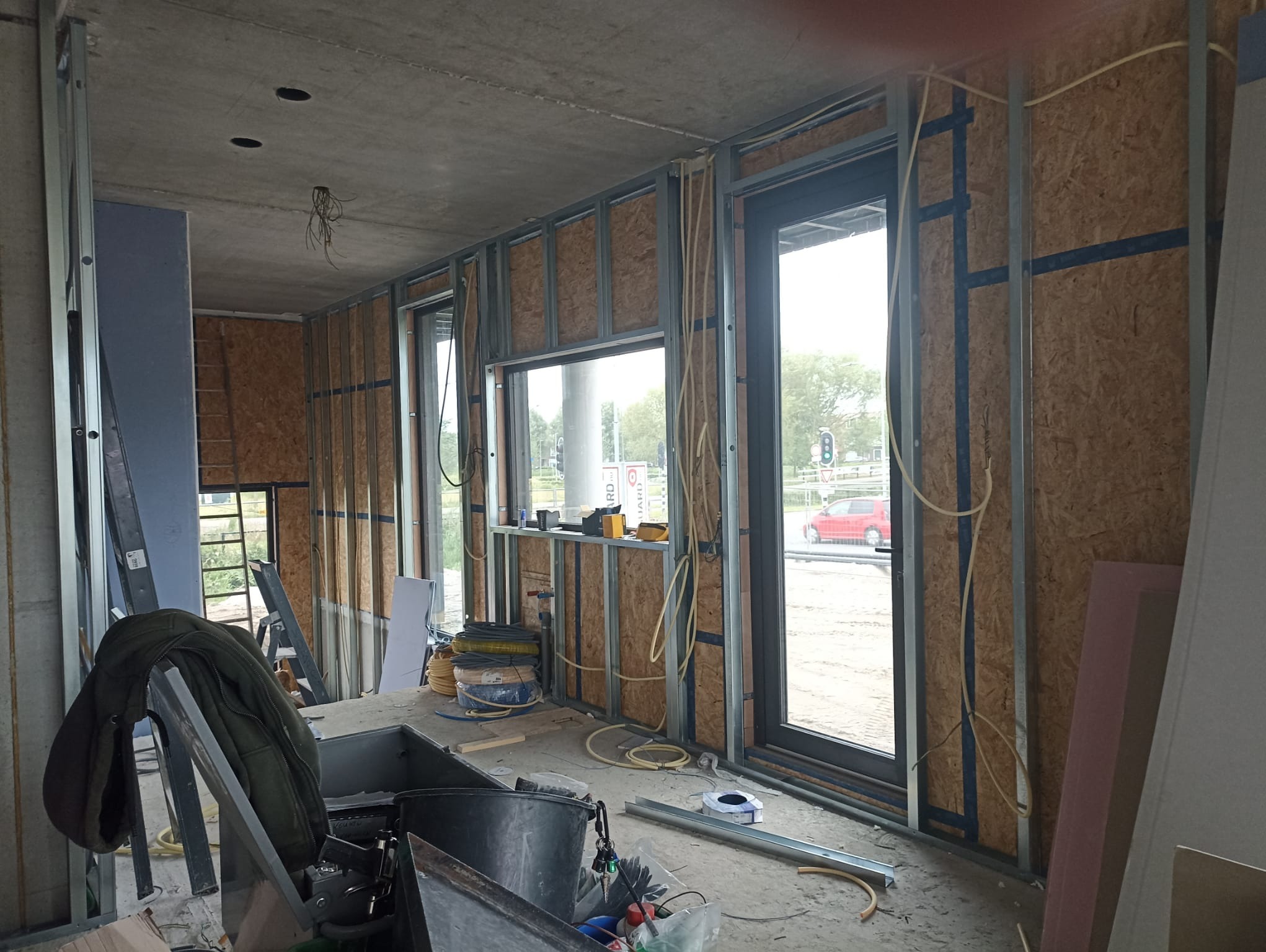 Wnętrze w trakcie prac wykończeniowych: metalowe profile, płyty OSB, instalacje elektryczne. Widoczne okna i narzędzia. Surowy stan, przygotowanie do dalszych prac.