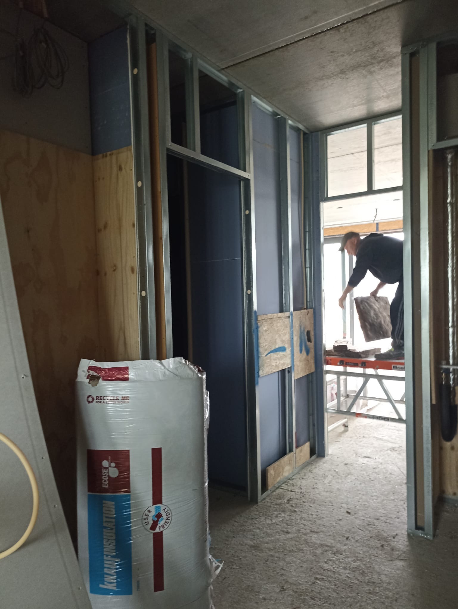 Montaż ścian działowych z płyty karton-gips na metalowym stelażu. Widoczna izolacja Knauf Insulation i pracownik układający materiał izolacyjny.