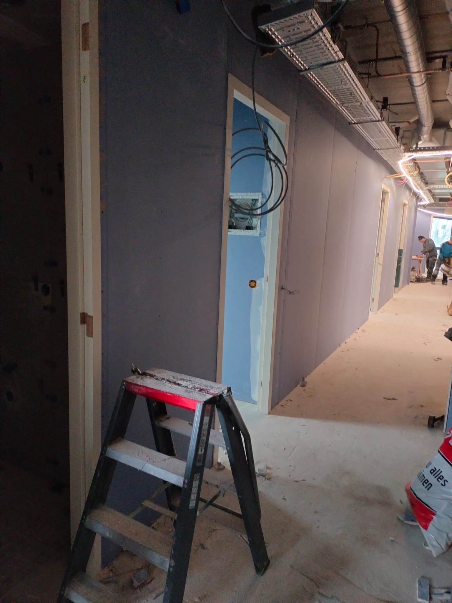 Korytarz w trakcie wykańczania płytami gipsowo-kartonowymi, widoczna drabina i instalacje pod sufitem. Ściany pomalowane na szaro-fioletowy kolor.