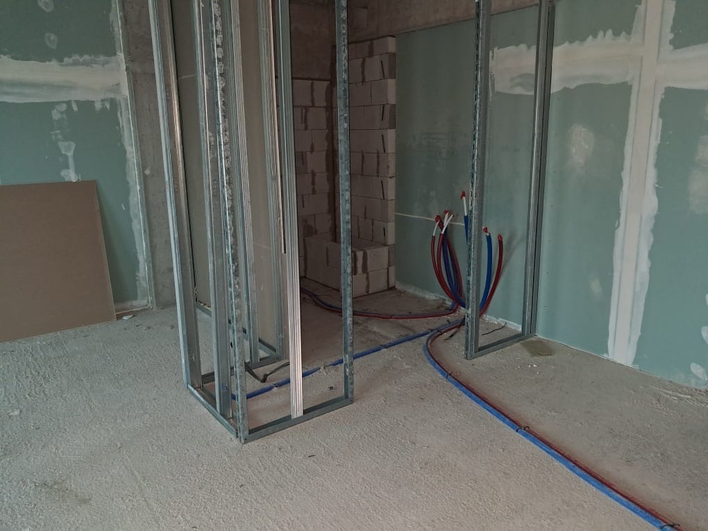 Wnętrze w trakcie remontu: metalowe profile pod płyty gipsowe, instalacja wodna w podłodze, ściany częściowo pokryte gładzią. Widoczny fragment ściany z pustaków.