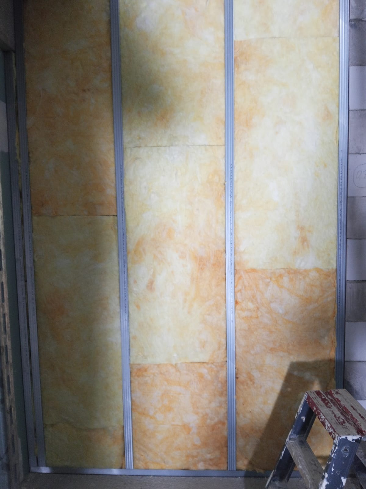 Montaż izolacji z wełny mineralnej w metalowym stelażu, widoczna drabina malarska. Prace wykończeniowe wewnątrz budynku, oświetlenie dzienne.