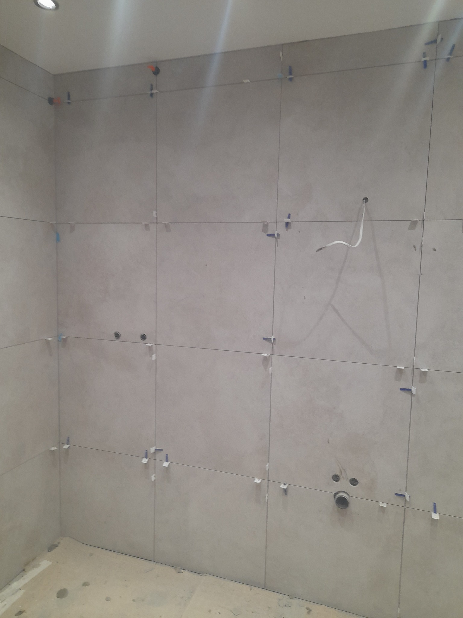 Ściana w trakcie układania płytek ceramicznych w łazience. Widoczne krzyżyki dystansowe i otwory na instalacje. Szare, matowe płytki ułożone równo.