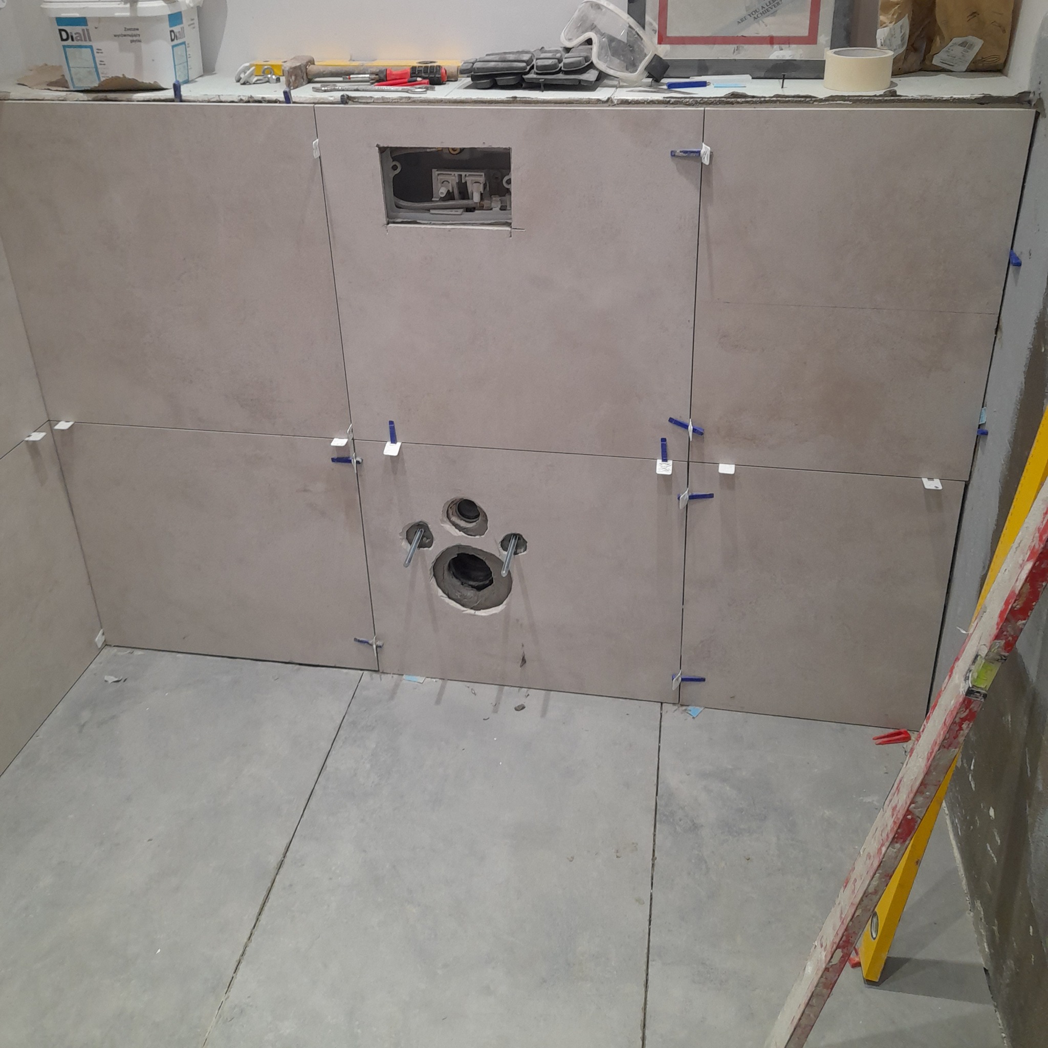 Ściana w trakcie układania płytek ceramicznych w łazience. Widoczne otwory na instalacje, poziomica i narzędzia glazurnicze. Użyto systemu poziomowania płytek.