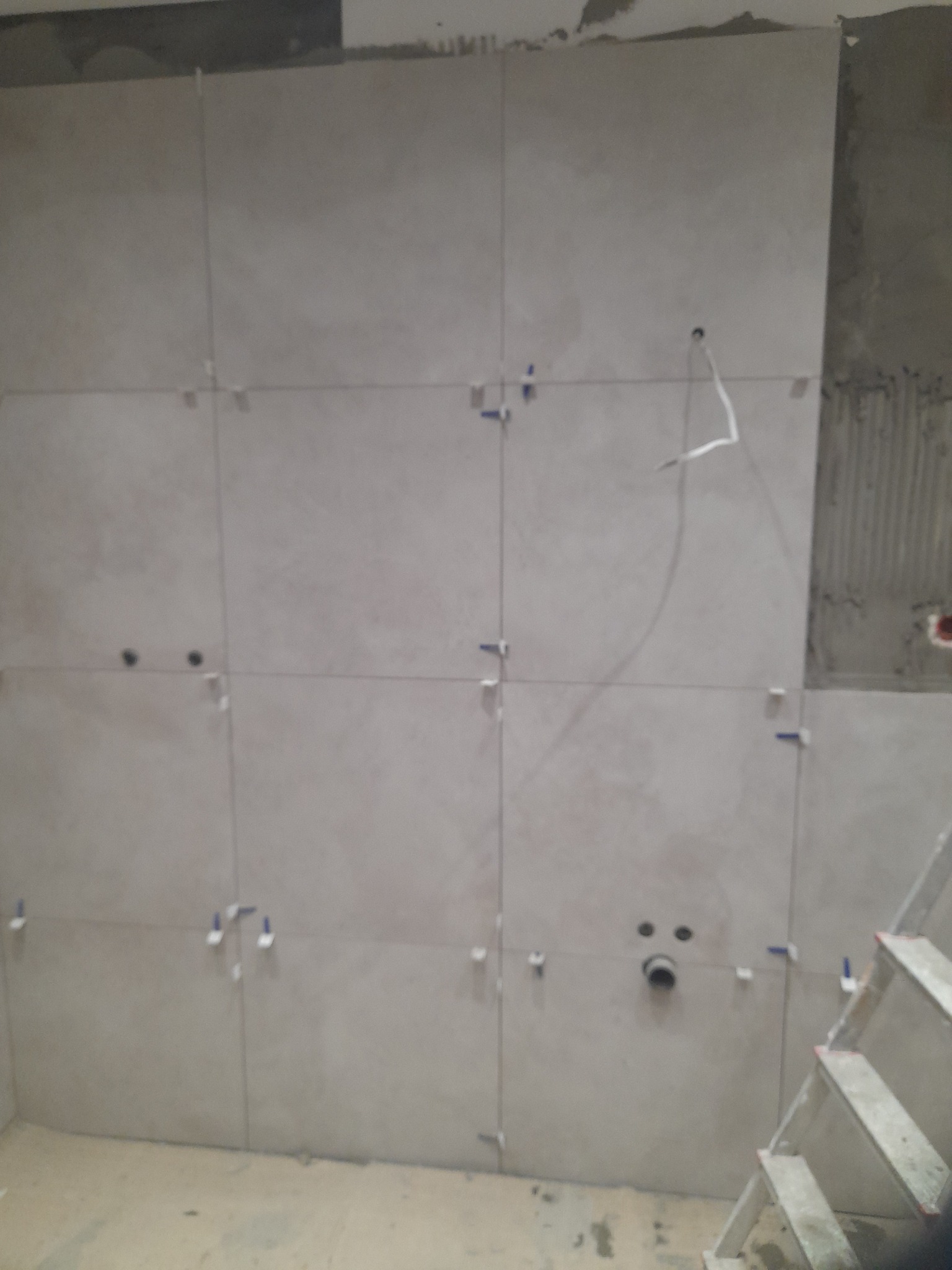 Ściana z jasnych, kwadratowych płytek, częściowo ułożonych, z widocznymi krzyżykami dystansowymi i fragmentem drabiny. Widoczne otwory na instalacje.