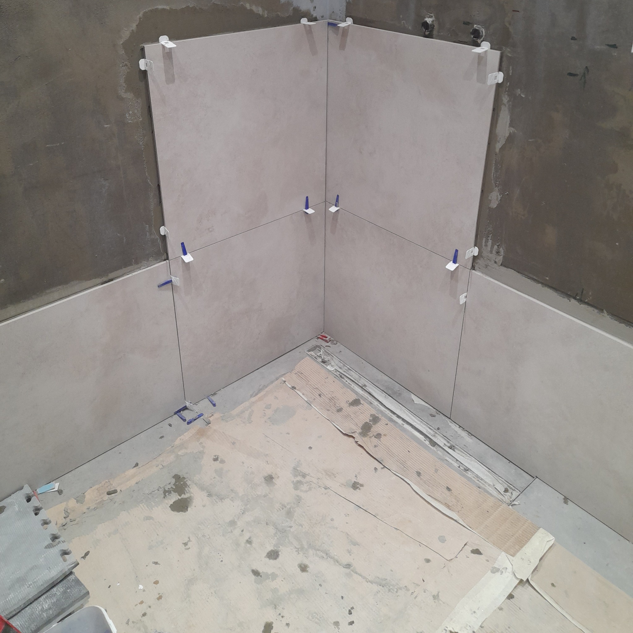 Układanie jasnych płytek w rogu pomieszczenia, widoczne niebieskie krzyżyki dystansowe i sznurki poziomujące. Surowe ściany w tle, przygotowanie do dalszych prac.