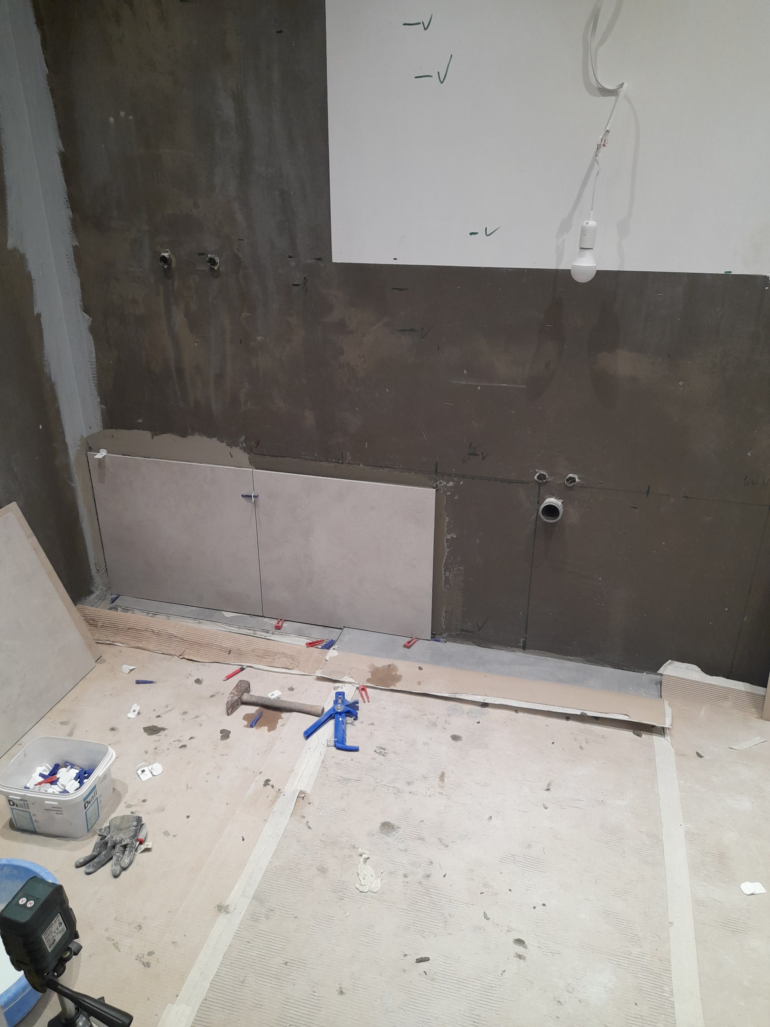 Fragment ściany w trakcie układania płytek w łazience. Widoczne narzędzia glazurnicze, poziomica laserowa i częściowo ułożone płytki w jasnym odcieniu.