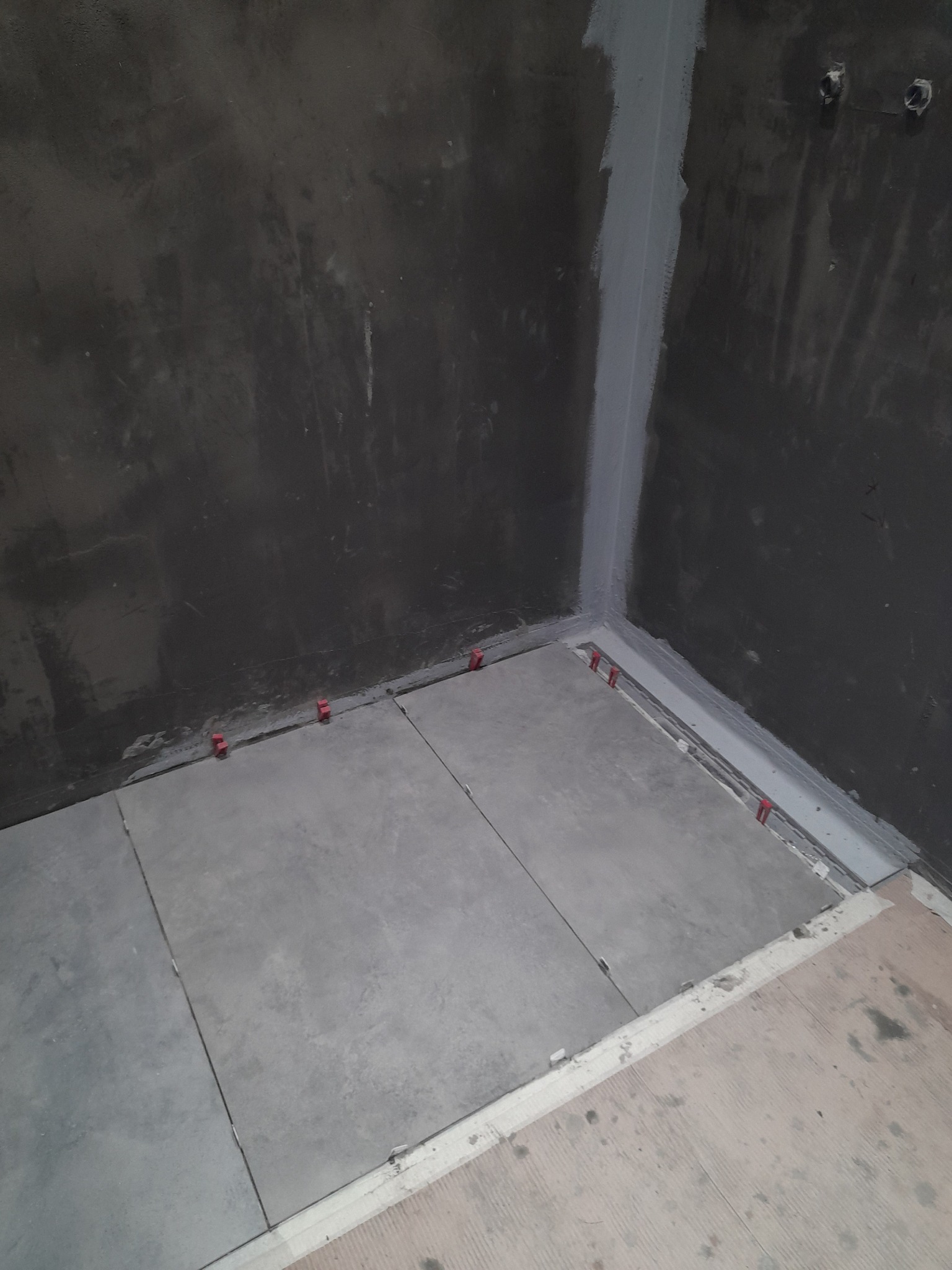 Szare płytki podłogowe w trakcie układania w rogu pomieszczenia. Widoczne czerwone klipsy poziomujące i szara hydroizolacja na styku ściany i podłogi.