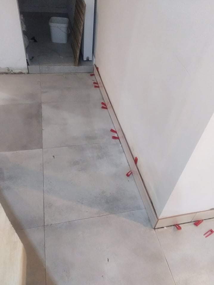 Ułożone szare płytki podłogowe z czerwonymi klipsami poziomującymi przy ścianie. Widoczny fragment progu i ściany. Ujęcie z góry, blisko podłogi.