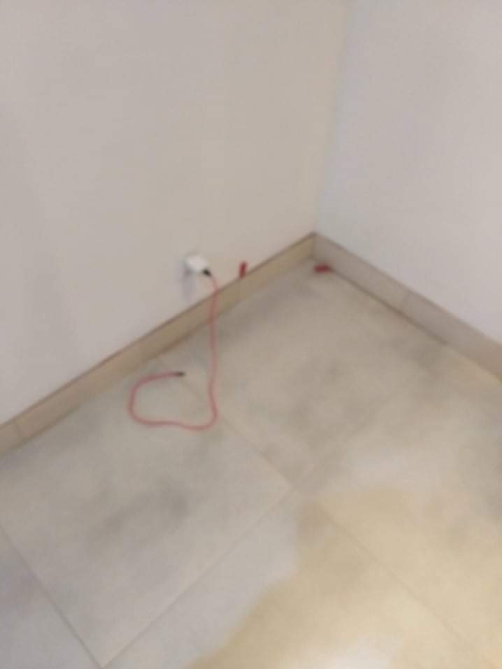 Białe płytki podłogowe w rogu pokoju, widoczna listwa przypodłogowa i kabel zasilający przy ścianie. Minimalistyczne wnętrze w trakcie wykańczania.