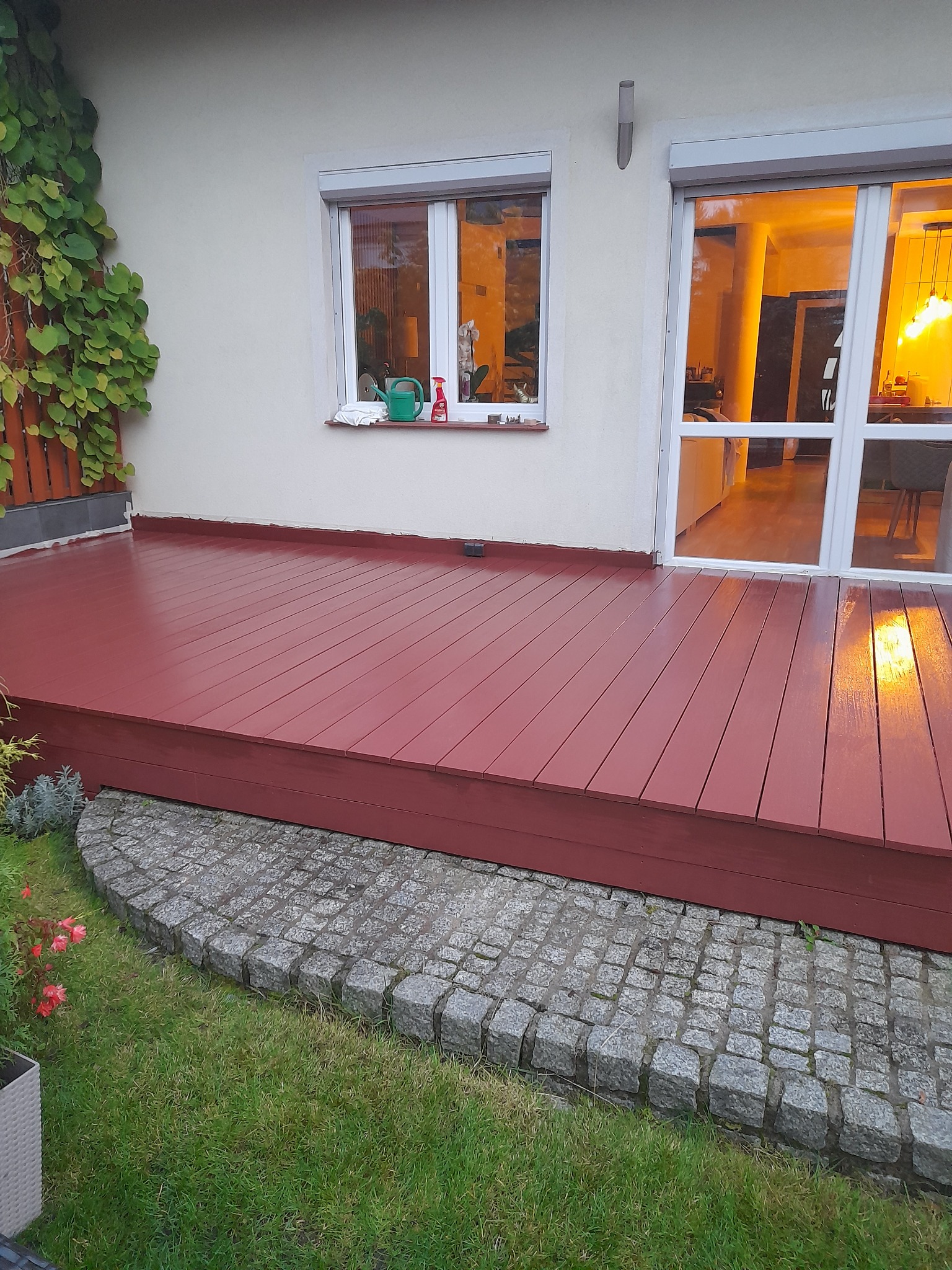 Czerwony, drewniany taras przy domu z oknem i roślinami. Widoczne kostki brukowe i trawa. W oknie odbija się ciepłe światło z wnętrza domu.