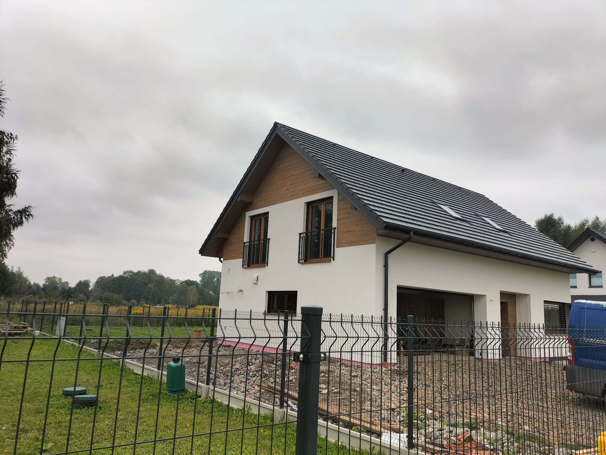 Nowoczesny dom jednorodzinny z białymi ścianami i ciemną dachówką, widoczny zza ogrodzenia. Uwagę zwraca drewniana elewacja pod dachem i teren w trakcie zagospodarowywania.