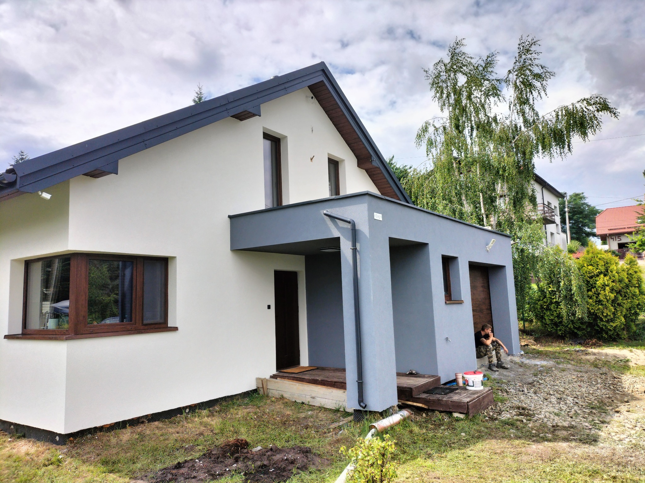 Nowoczesny dom po renowacji elewacji w odcieniach bieli i szarości. Widoczny pracownik siedzący przed wejściem, farby i narzędzia. Ujęcie z poziomu gruntu.