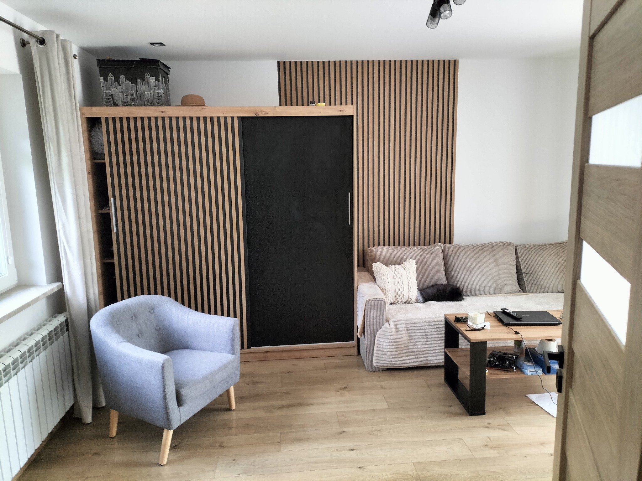 Nowoczesny salon z drewnianą ścianą z lameli, szarą sofą i fotelem. Na ścianie szafa z czarnymi przesuwnymi drzwiami. Na podłodze jasne panele. Widok zza drzwi.