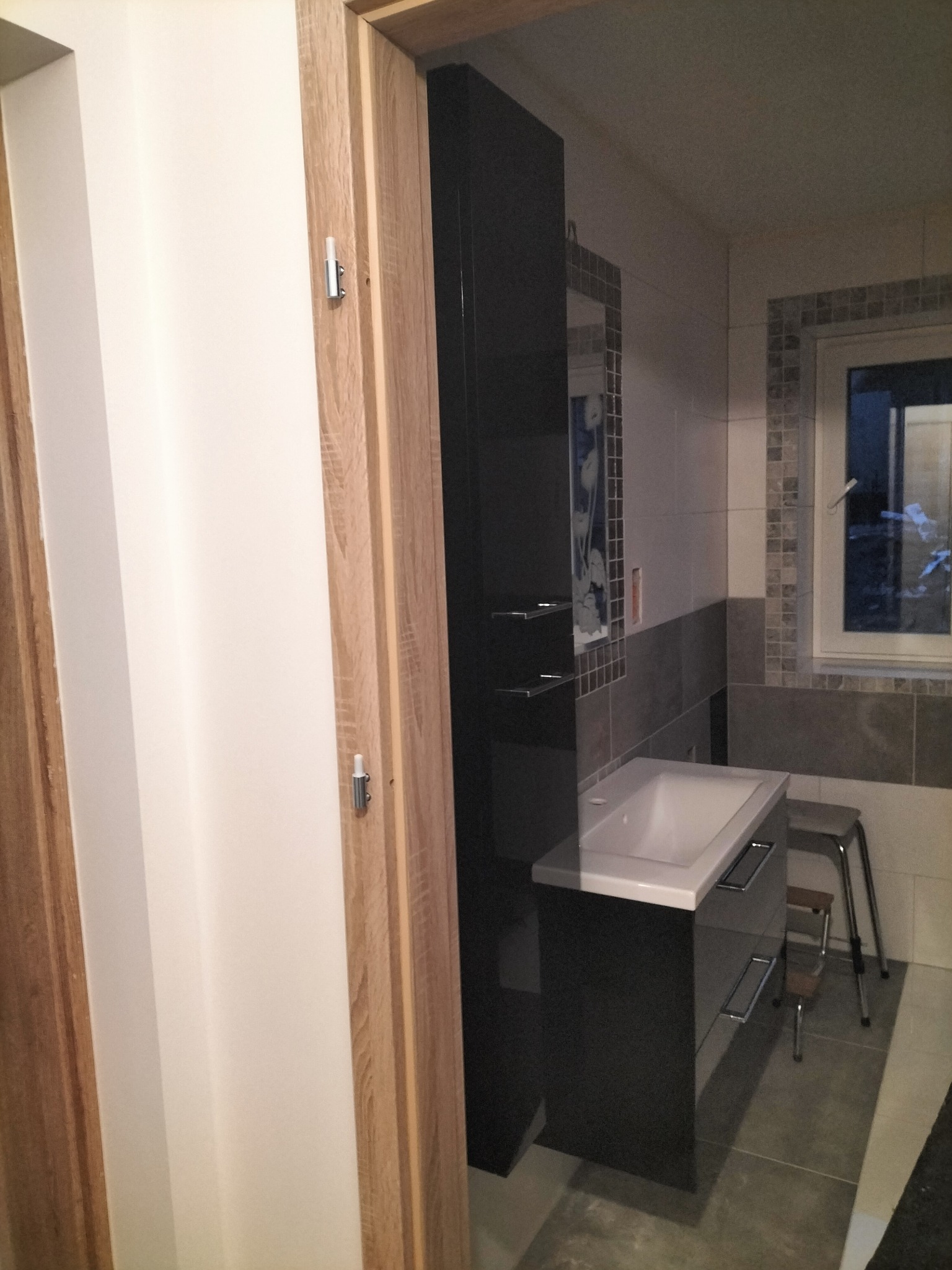 Nowoczesna łazienka z czarną szafką pod umywalką, lustrem w mozaikowej ramie i oknem. Widoczny fragment drewnianych drzwi. Stonowana kolorystyka płytek.