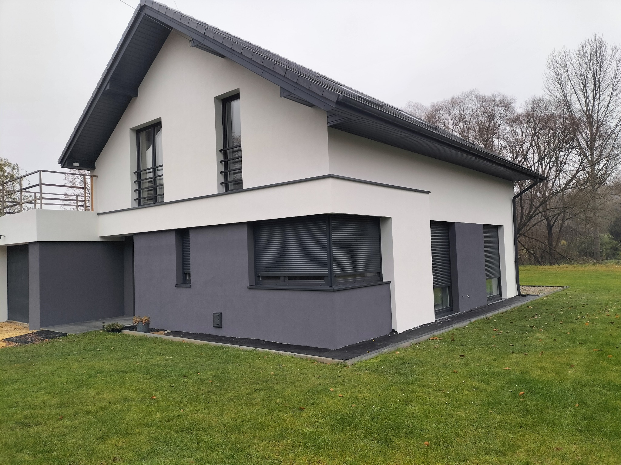 Dwukondygnacyjny dom z szarą elewacją dolną i białą górną, z ciemnymi roletami okiennymi i grafitowym dachem, otoczony zielonym trawnikiem w pochmurny dzień.