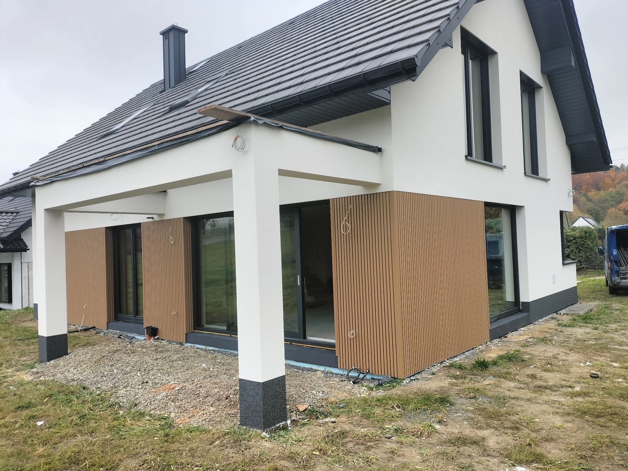 Nowoczesny dom jednorodzinny z drewnianą elewacją i dużymi oknami przesuwnymi. Widoczne detale wykończenia i otoczenie domu w trakcie prac.