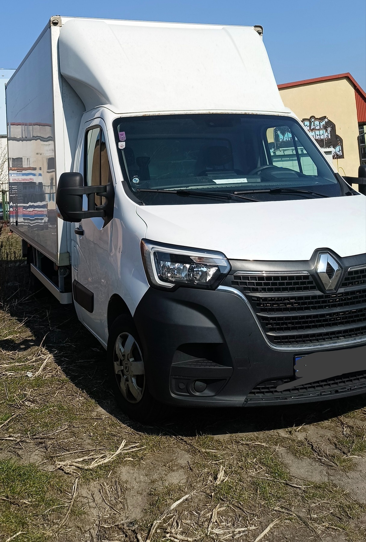 Biały samochód dostawczy Renault Master z wysoką zabudową, zaparkowany na trawiastym terenie w słoneczny dzień. Widoczny przód pojazdu i fragment boku.