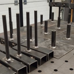 ATS Sowa - Spawane elementy metalowe: gwintowane pręty i rury przymocowane do kwadratowych płyt na perforowanym stole spawalniczym. Widoczne ślady spawania.