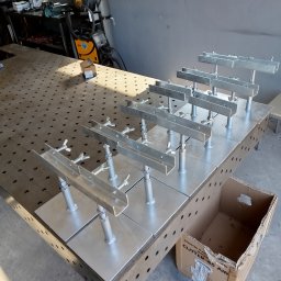 ATS Sowa - Metalowe elementy konstrukcyjne, prawdopodobnie spawane, ustawione rzędami na perforowanym stole warsztatowym w szarym pomieszczeniu. Widoczny karton i sprzęt spawalniczy w tle.