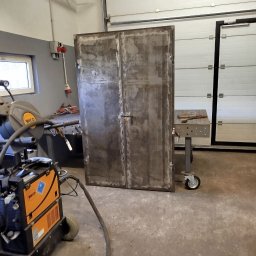 ATS Sowa - Surowe, metalowe dwuskrzydłowe drzwi w warsztacie, obok spawarka WECO i stół spawalniczy na kółkach. Widoczne ślady obróbki metalu.