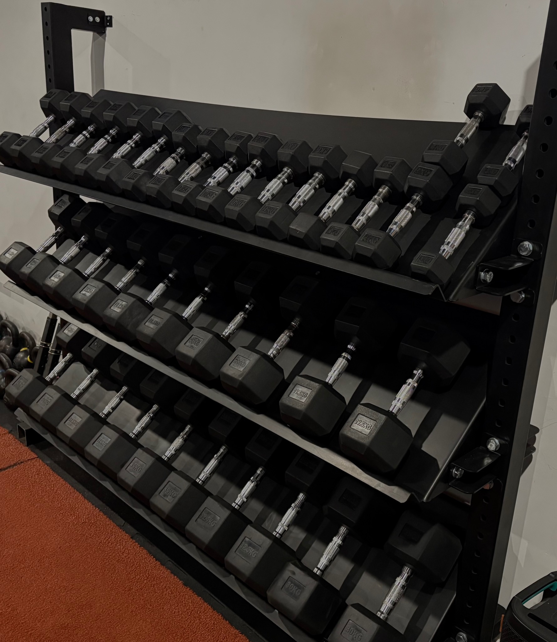Stojak z kompletem czarnych hantli sześciokątnych o różnych wagach, ułożonych rzędami na trzech poziomach. Widoczna podłoga w kolorze ceglastym.