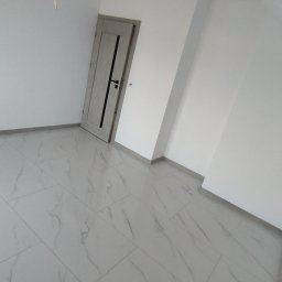Glazura usługi ogólnobudowlane - Jasne wnętrze po remoncie: białe ściany, podłoga z płytek imitujących marmur, szare drzwi z czarną klamką. Widoczny fragment kabla wystający z sufitu.