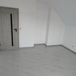 Glazura usługi ogólnobudowlane - Jasne wnętrze po remoncie: białe ściany, podłoga z płytek imitujących marmur, szare drzwi z czarnym akcentem. Widoczny fragment kabla zwisającego z sufitu.