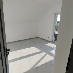 Glazura usługi ogólnobudowlane - Jasne wnętrze poddasza z białymi ścianami i podłogą z płytek imitujących marmur, widziane zza uchylonych drzwi. Wpadające światło słoneczne podkreśla wzór płytek.