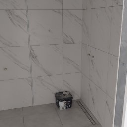 Glazura usługi ogólnobudowlane - Wykończona ściana łazienki z białych płytek imitujących marmur. W rogu stoi wiadro z farbą akrylową. Widoczny odpływ liniowy w podłodze.