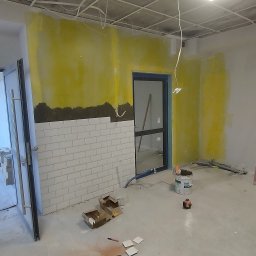 Glazura usługi ogólnobudowlane - Wnętrze w trakcie prac: fragment ściany z ułożonymi białymi płytkami, żółte ślady po malowaniu, narzędzia i materiały budowlane na podłodze. Widoczne kable pod sufitem.