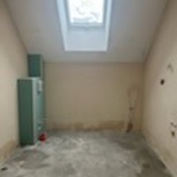 Glazura usługi ogólnobudowlane - Poddasze przed remontem: surowe ściany, betonowa podłoga, okno dachowe. Widoczne ślady wilgoci i niedokończone instalacje. Szafka w rogu pomieszczenia.