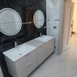 Glazura usługi ogólnobudowlane - Nowoczesna łazienka z podwójną umywalką na szafce w odcieniach beżu, okrągłe lustra i marmurowe płytki w czerni i bieli. Widok na otwarte drzwi.