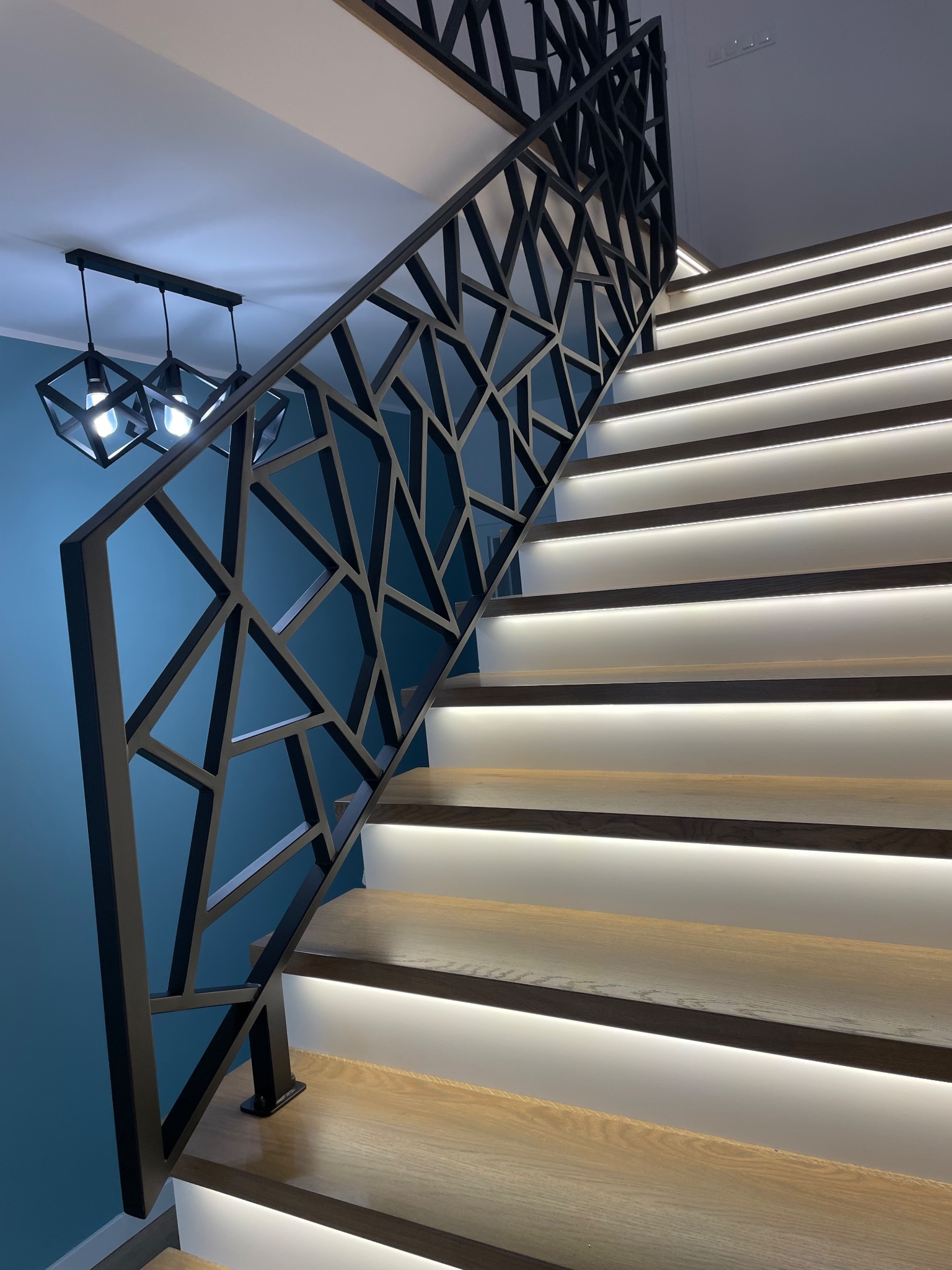 Nowoczesne schody z drewnianymi stopniami, podświetleniem LED i geometryczną, czarną balustradą metalową na tle turkusowej ściany. Widoczny fragment designerskiej lampy.