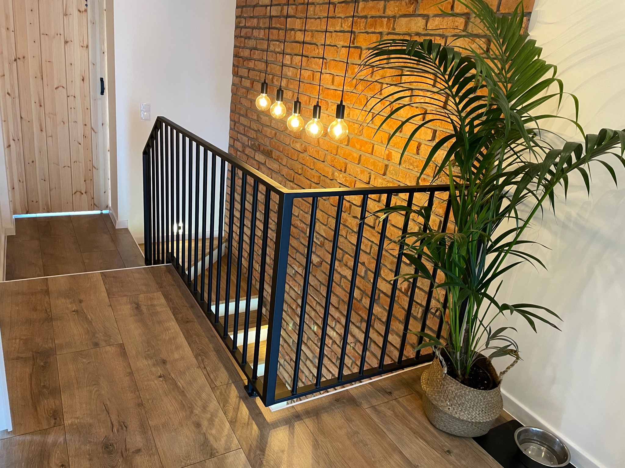 Nowoczesne schody z czarną, metalową balustradą na tle ceglanej ściany. Oświetlenie w stylu loft i roślina doniczkowa dodają wnętrzu charakteru.