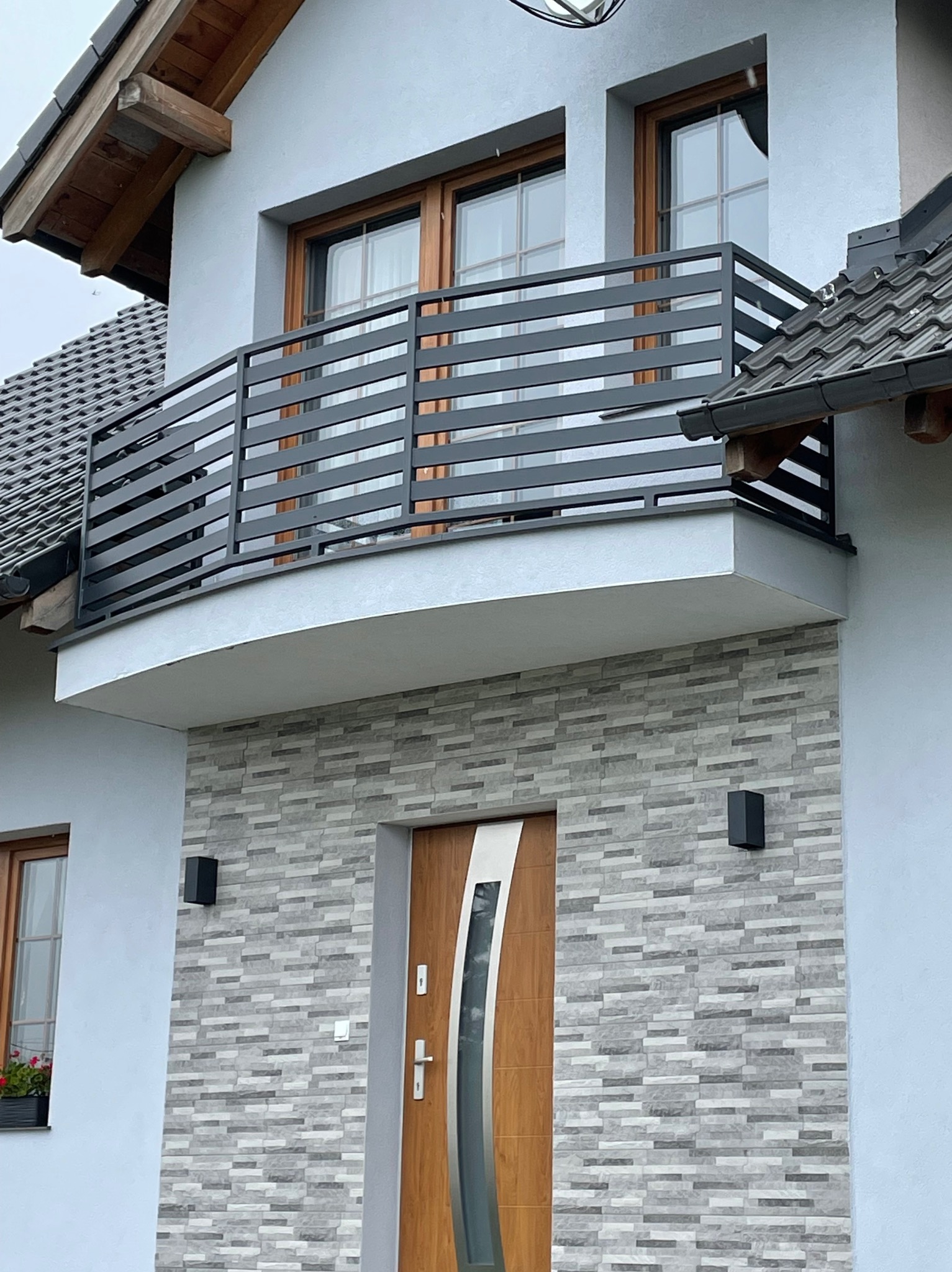 Nowoczesny balkon z szarą, poziomą balustradą metalową, drewniane okna i drzwi wejściowe z dekoracyjną okładziną kamienną na elewacji domu.