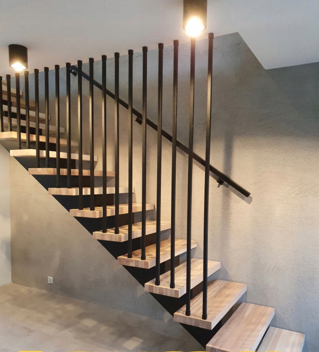 Nowoczesne schody z drewnianymi stopniami i czarną, metalową balustradą w minimalistycznym stylu. Oświetlenie punktowe podkreśla surowy charakter wnętrza.