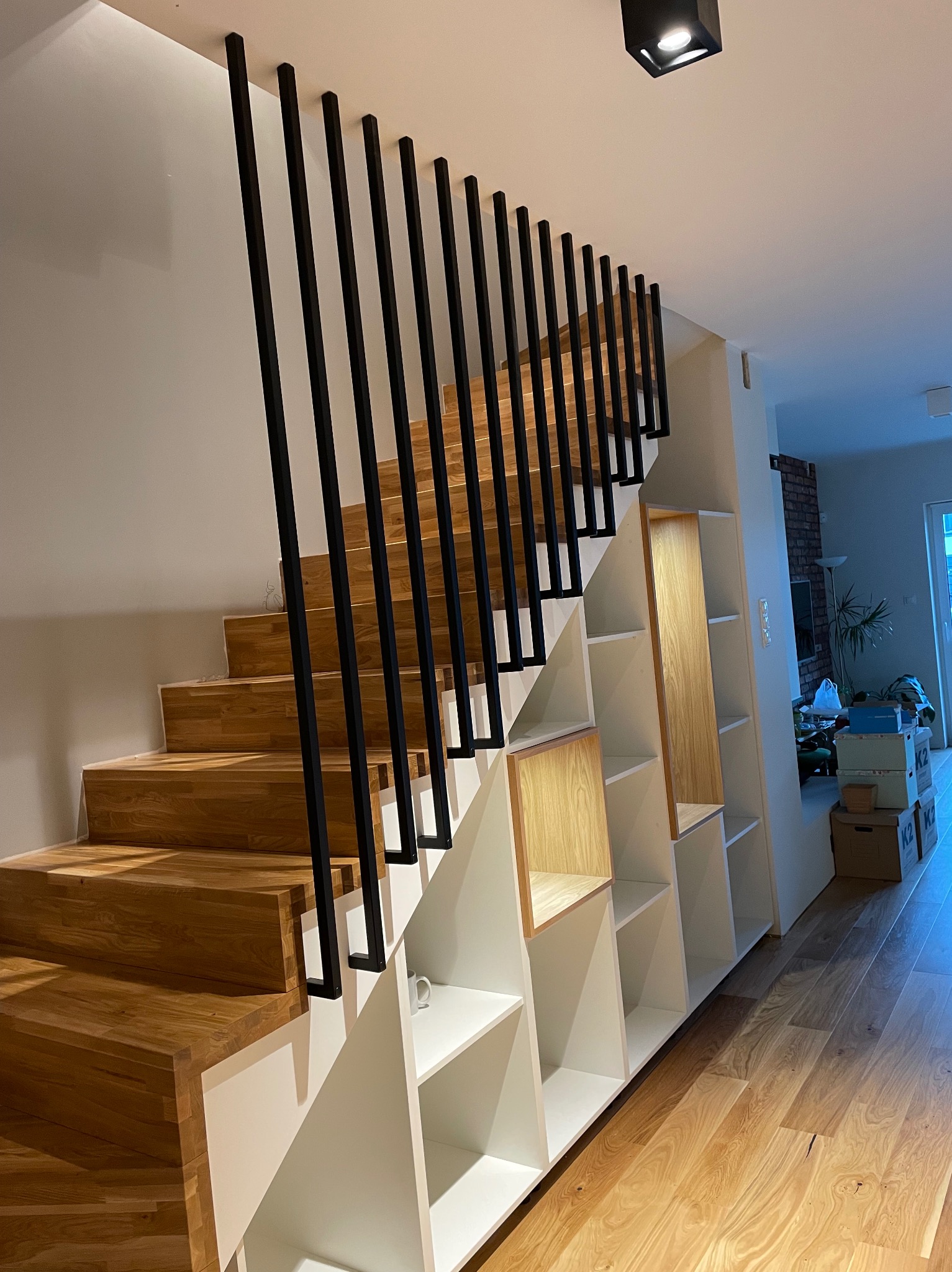 Drewniane schody z czarnymi, metalowymi poręczami i wbudowanymi białymi półkami, oświetlone punktowym światłem, w nowoczesnym wnętrzu domu.